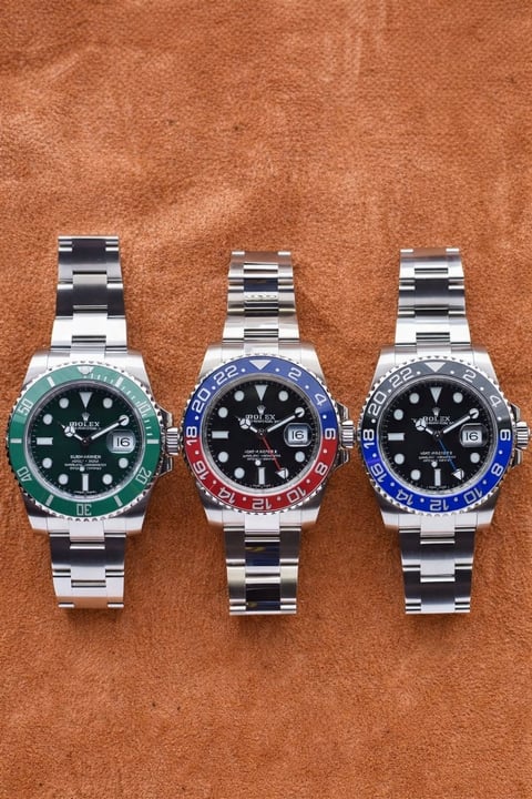 Three Rolex GMT-Master II watches with steel bracelets and colored bezels - green, red, and blue - displayed on tan leather background