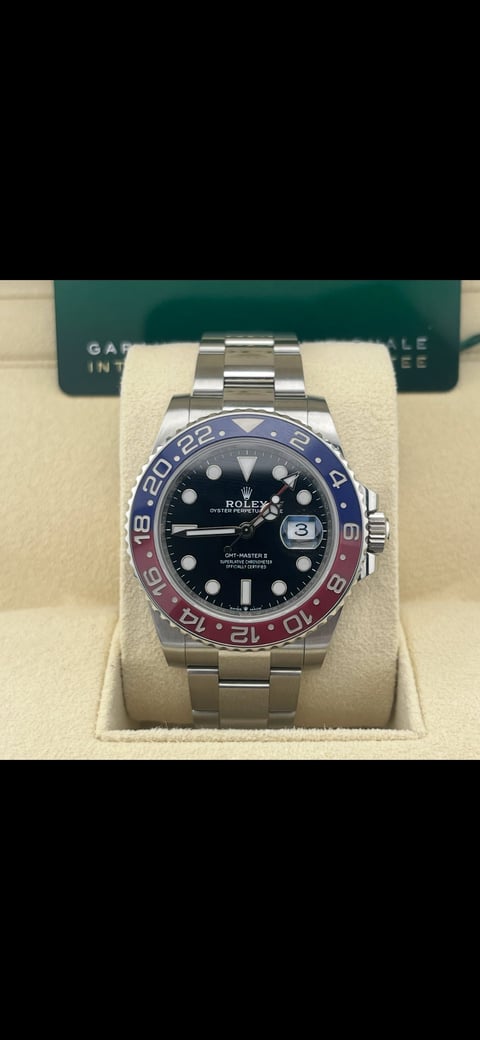 Rolex GMT-Master II watch with steel bracelet, black dial, and blue-red ceramic bezel, displayed on cream cushion with green Rolex box