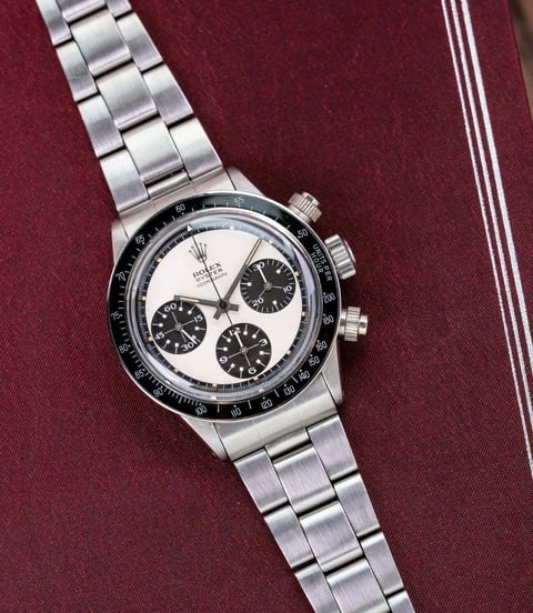 Steel chronograph watch with white dial and black subdials on matching metal bracelet against burgundy background