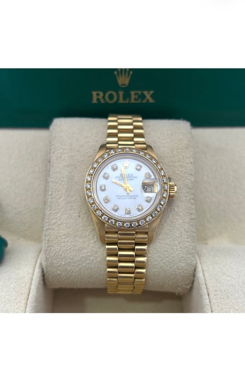 Gold Rolex watch with diamond bezel and white mother-of-pearl dial on cream display stand with green Rolex box